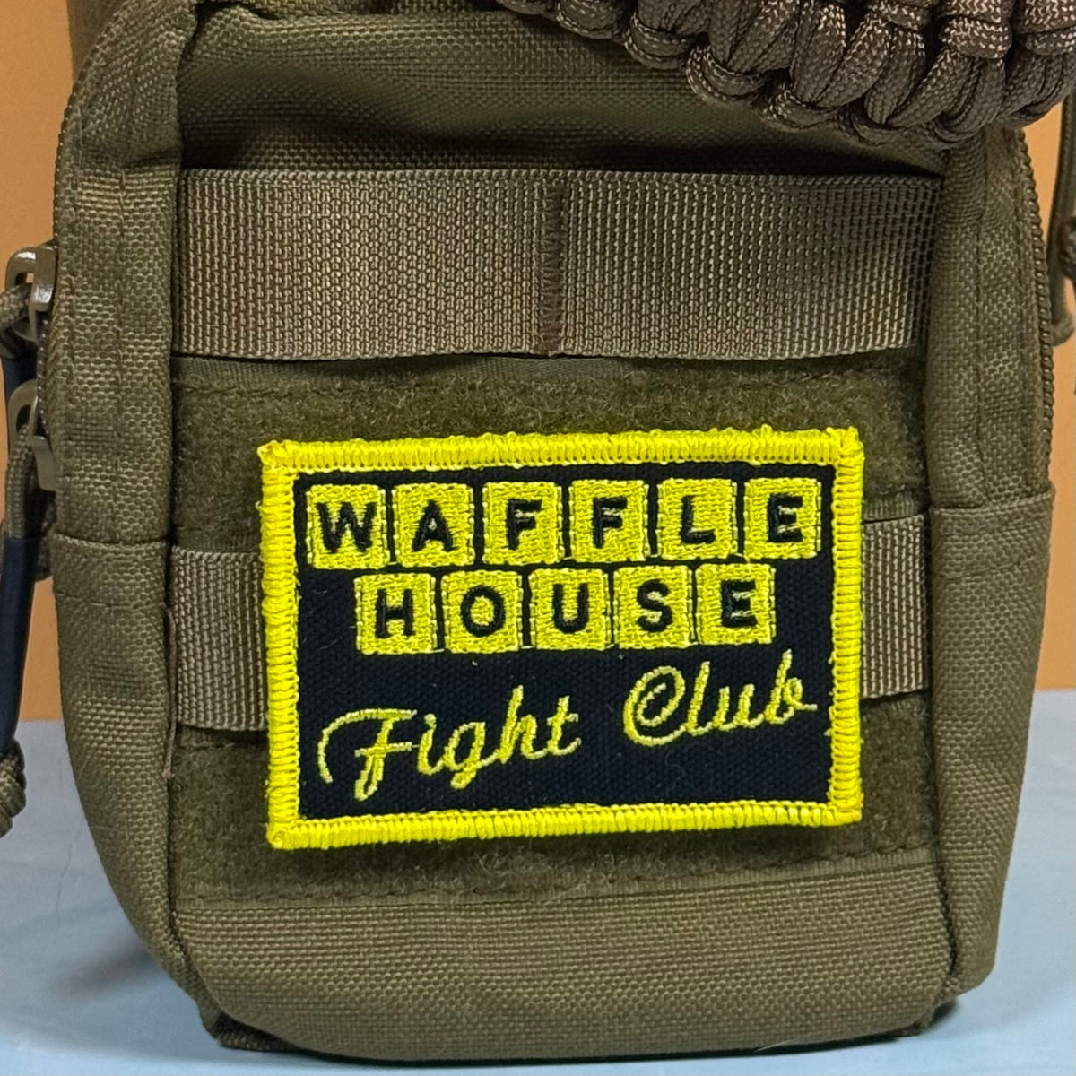 An olive green tactical pouch featuring a rectangular embroidered patch that reads 'WAFFLE HOUSE Fight Club' in yellow on a black background.