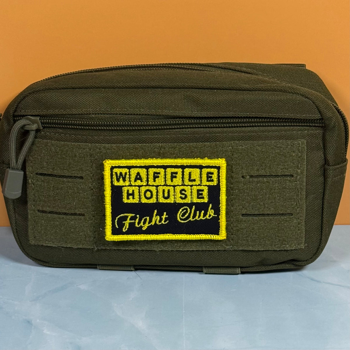 An olive green tactical pouch with a front zippered pocket and a prominent embroidered patch reading 'WAFFLE HOUSE Fight Club' in yellow and black, set against an orange background.