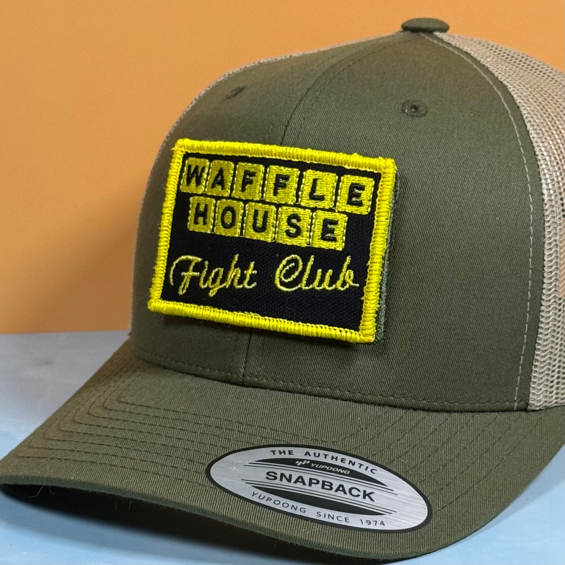 An olive green and beige trucker cap with a custom embroidered patch reading 'WAFFLE HOUSE Fight Club' in yellow and black. The cap features a structured front panel, olive green visor, and beige mesh back, with a visible Snapback sticker on the brim.