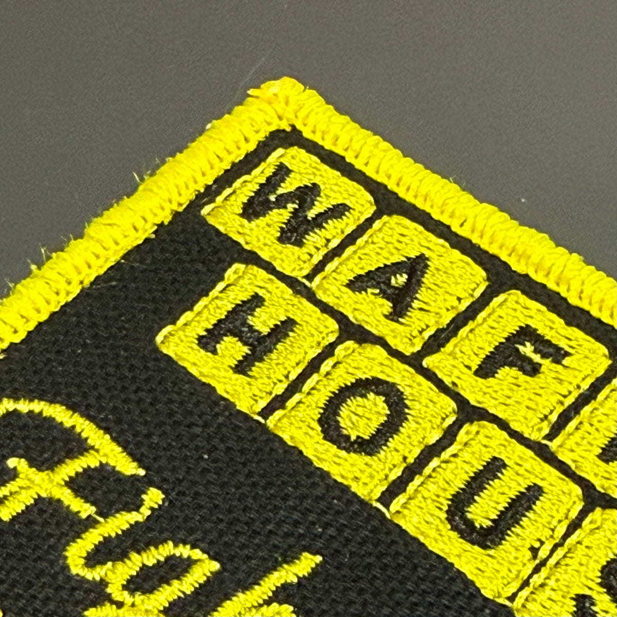 Close-up of a black embroidered patch with bright yellow border and yellow lettering on a dark gray background.