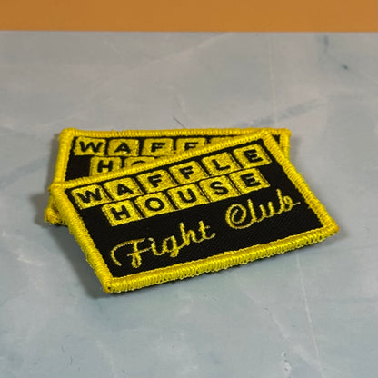 Two embroidered rectangular patches with yellow borders and black centers reading 'WAFFLE HOUSE Fight Club' on a light gray marble-like surface.