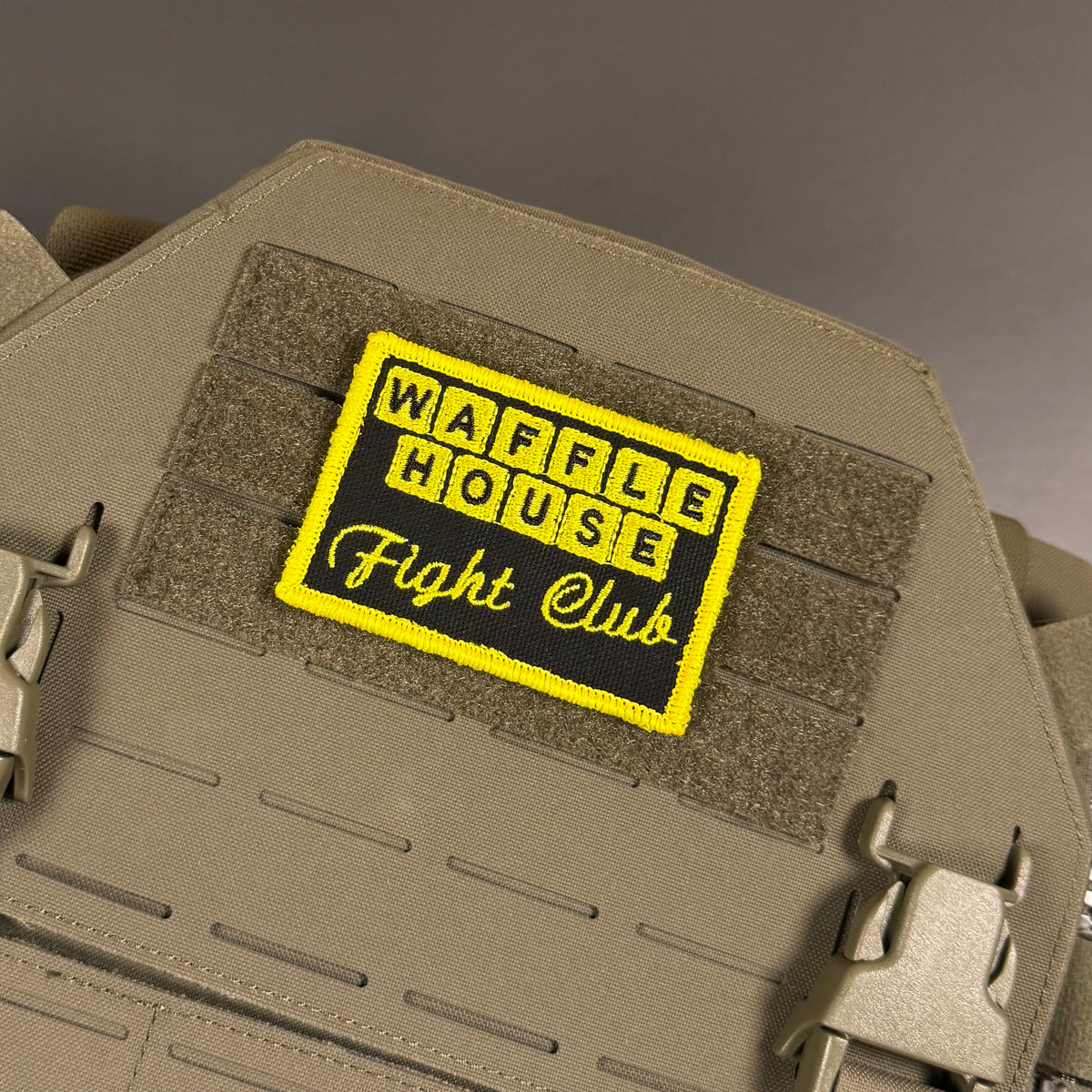 A rectangular embroidered patch reading 'WAFFLE HOUSE Fight Club' in yellow and black, attached to tan tactical gear with hook-and-loop panels and metal quick-release clips.