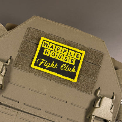 A rectangular embroidered patch reading 'WAFFLE HOUSE Fight Club' in yellow and black, attached to tan tactical gear with hook-and-loop panels and metal quick-release clips.