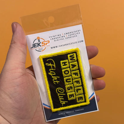 A hand holds a packaged embroidered patch that reads 'WAFFLE HOUSE Fight Club' in yellow on a black background, with a clear plastic sleeve and branded backing card against an orange backdrop.