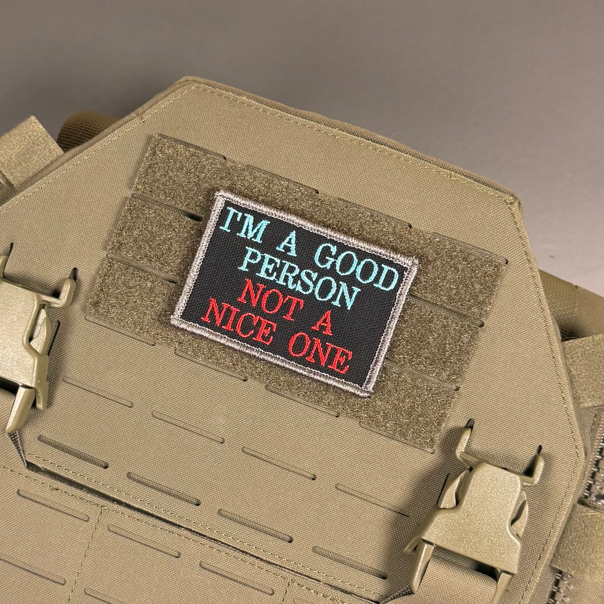 A rectangular embroidered tactical patch with the text 'I'M A GOOD PERSON NOT A NICE ONE' in light blue and red lettering on a black background, shown attached to tan coyote brown MOLLE webbing on a tactical vest or plate carrier.