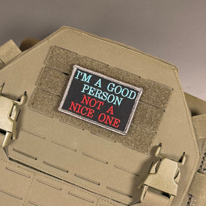 A rectangular embroidered tactical patch with the text 'I'M A GOOD PERSON NOT A NICE ONE' in light blue and red lettering on a black background, shown attached to tan coyote brown MOLLE webbing on a tactical vest or plate carrier.