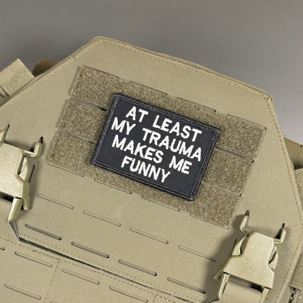 A rectangular black tactical patch with white embroidered text reading 'AT LEAST MY TRAUMA MAKES ME FUNNY' attached to a tan coyote brown MOLLE plate carrier vest.