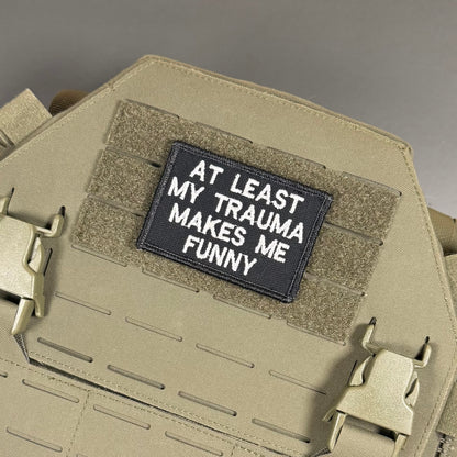 A rectangular black tactical patch with white embroidered text reading 'AT LEAST MY TRAUMA MAKES ME FUNNY' attached to a tan coyote brown MOLLE plate carrier vest.