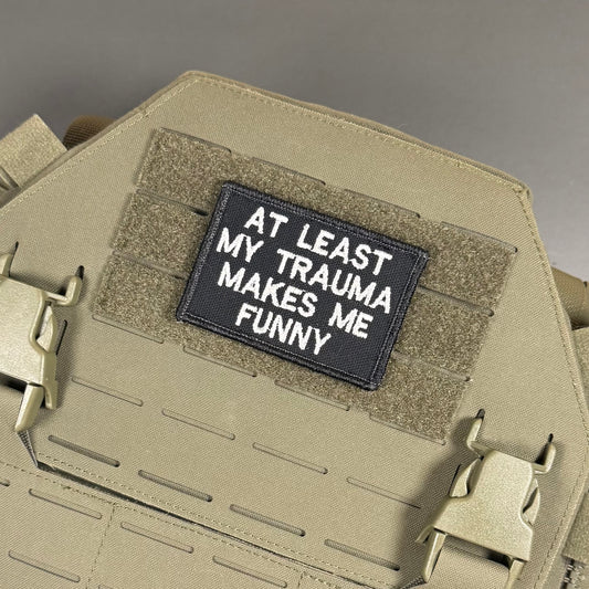 At Least My Trauma Makes Me embroidered morale patch: black tactical patch with white embroidered text reading 'AT LEAST MY TRAUMA MAKES ME FUNNY' attached to a tan coyote brown MOLLE plate carrier vest.