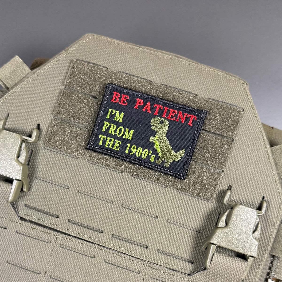 A rectangular embroidered patch with the text 'BE PATIENT I'M FROM THE 1900's' in red and yellow on a black background, attached to a tan tactical plate carrier.