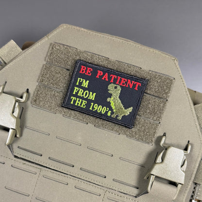 A rectangular embroidered patch with the text 'BE PATIENT I'M FROM THE 1900's' in red and yellow on a black background, attached to a tan tactical plate carrier.