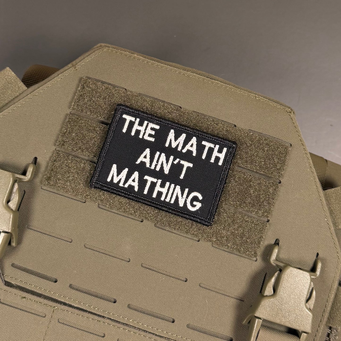 A rectangular black tactical patch with white embroidered text reading 'THE MATH AIN'T MATHING' attached to tan MOLLE webbing on a tan tactical vest or plate carrier.
