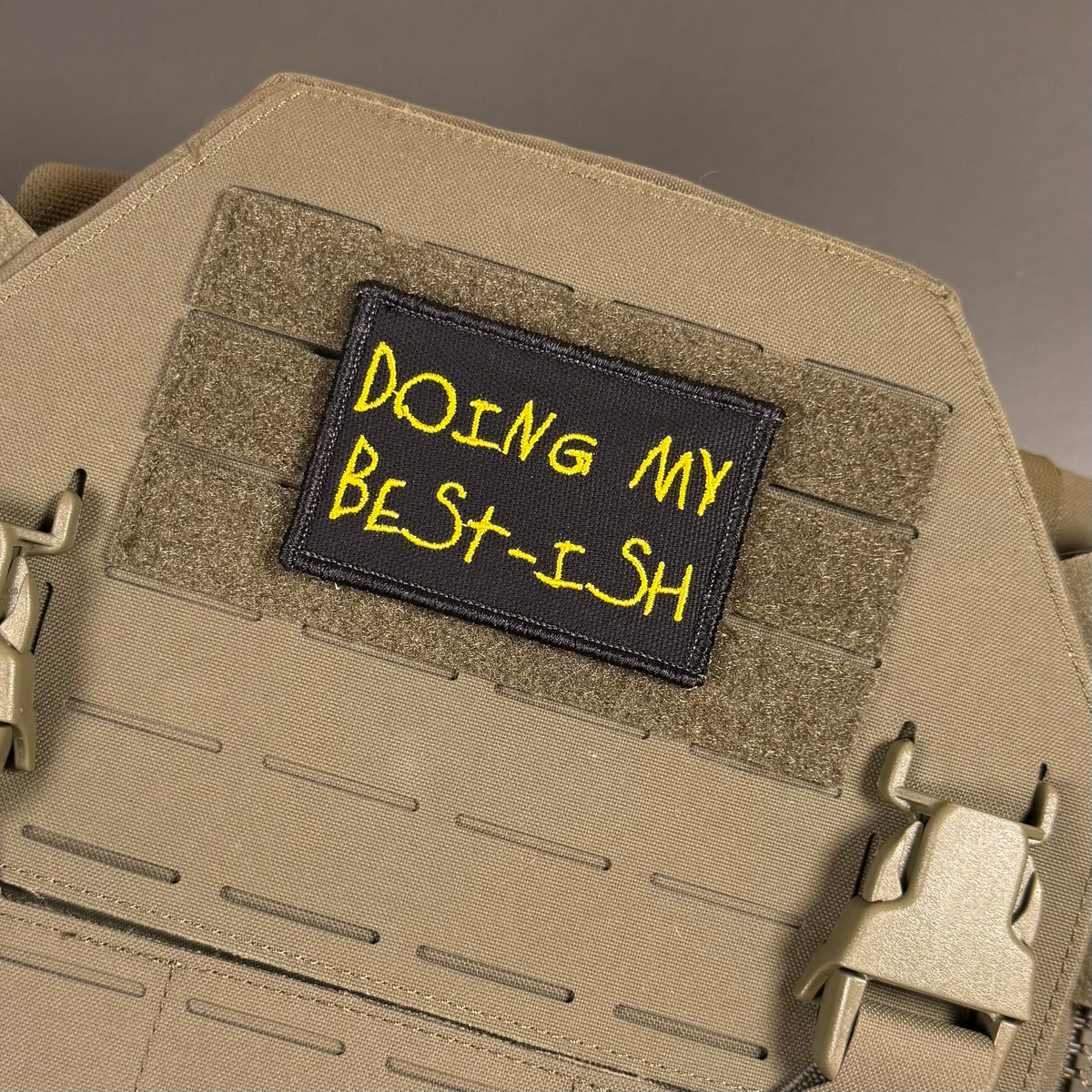 A rectangular black tactical patch with yellow embroidered text reading 'DOING MY BEST-ISH' attached to tan webbing on a tan tactical vest or plate carrier.