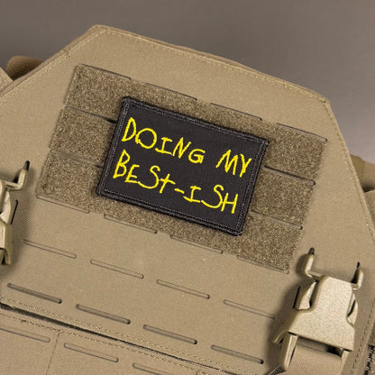 A rectangular black tactical patch with yellow embroidered text reading 'DOING MY BEST-ISH' attached to tan webbing on a tan tactical vest or plate carrier.