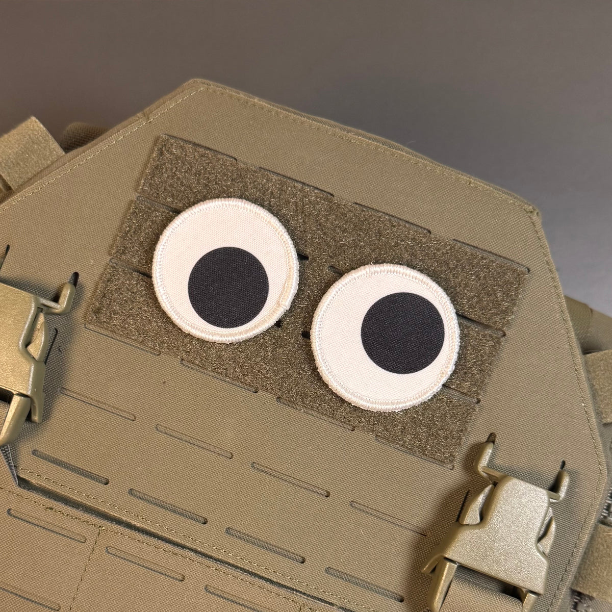 A close-up of a tan tactical vest or plate carrier with two circular googly-style eyes attached on a hook-and-loop panel.