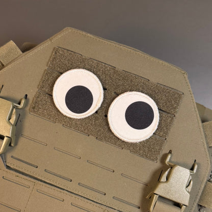 A close-up of a tan tactical vest or plate carrier with two circular googly-style eyes attached on a hook-and-loop panel.