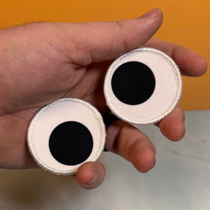 A hand holding two round white fabric patches with black circular centers, photographed against a split orange and light gray background.