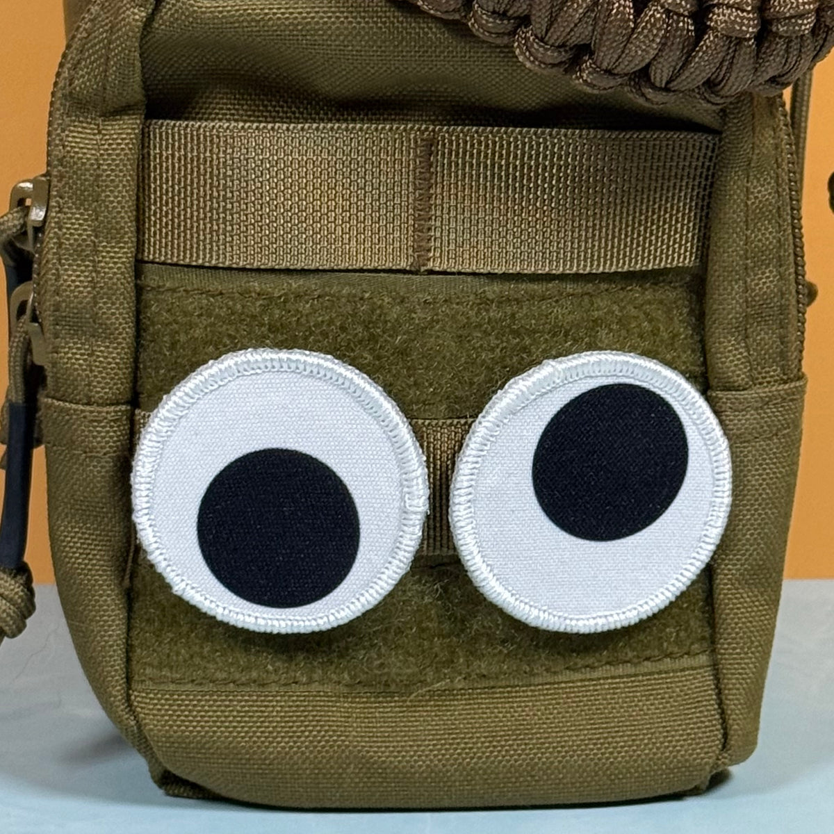 An olive green tactical pouch featuring a cute cartoon face made from two white circular patches with black centers, resembling big eyes.