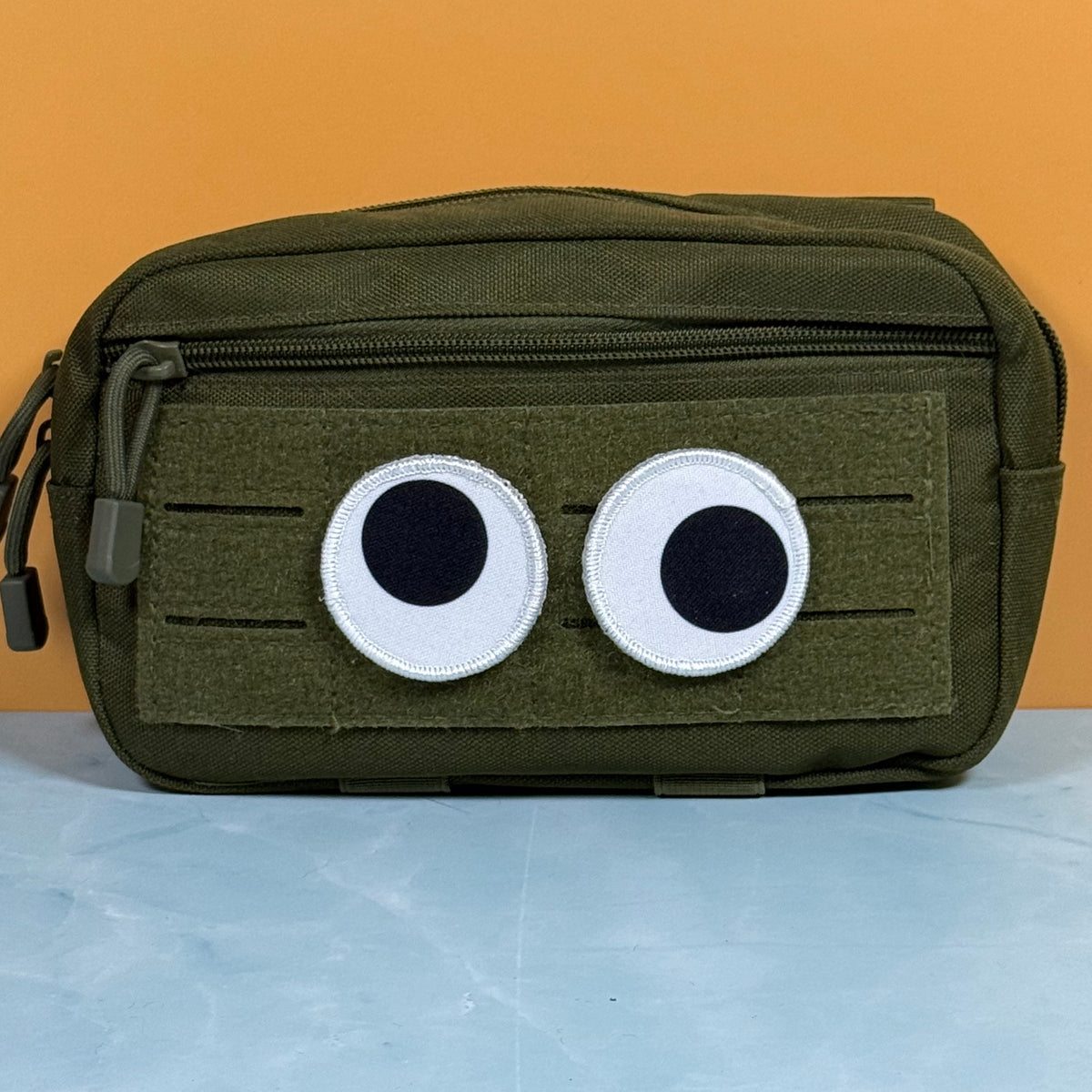 An olive green tactical pouch with a front zipper and a patch of two cartoon eyes on a hook-and-loop panel, set against an orange background and light blue surface.