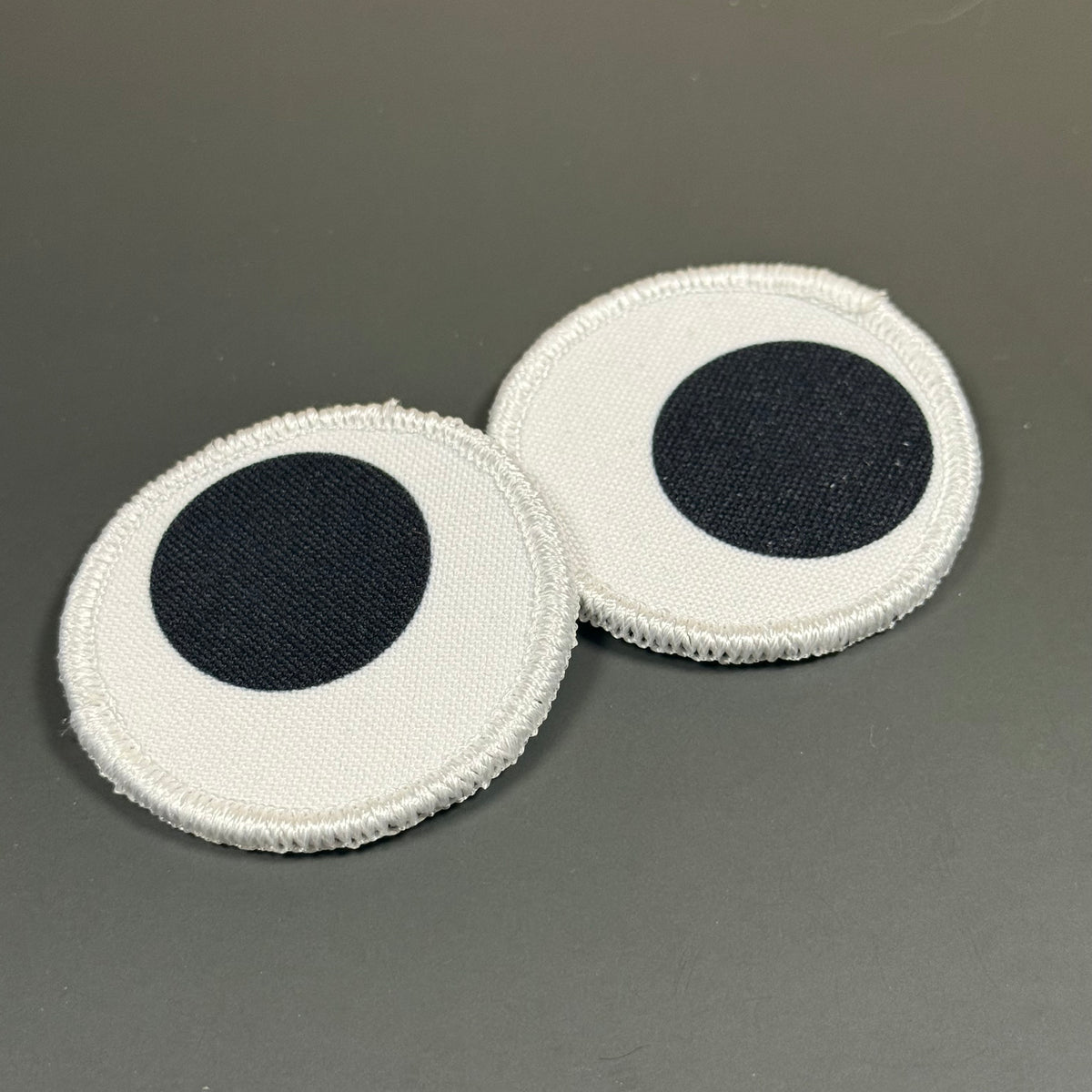 Two round white fabric patches with a black circular center, resembling eyes, shown on a dark gray background.