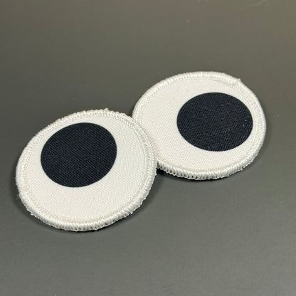 Two round white fabric patches with a black circular center, resembling eyes, shown on a dark gray background.