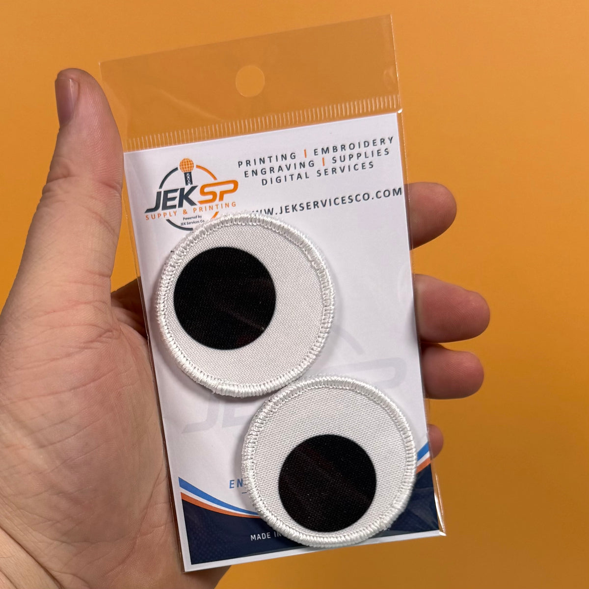 A hand holds a clear plastic package containing two circular embroidered patches with white stitched borders and black centers, against a solid orange background. The packaging card shows the brand name JEKSP.