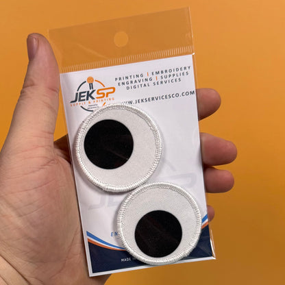 A hand holds a clear plastic package containing two circular embroidered patches with white stitched borders and black centers, against a solid orange background. The packaging card shows the brand name JEKSP.