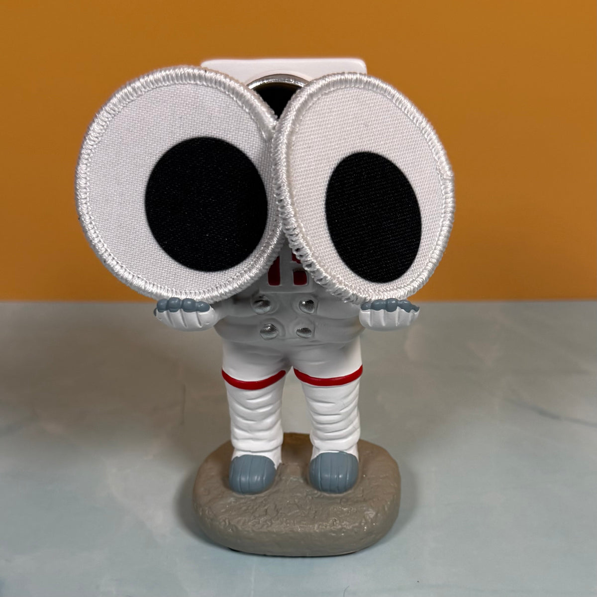 A novelty astronaut figurine with oversized white googly-style eyes, a white spacesuit, red stripe details, and a gray rock base, photographed against a two-tone orange and gray background.
