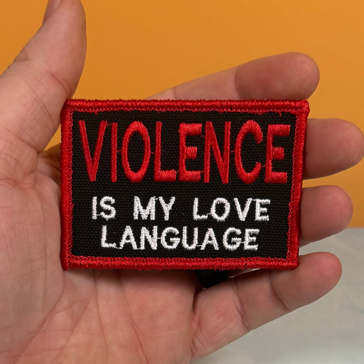 A rectangular embroidered patch held in a hand, featuring the text 'VIOLENCE IS MY LOVE LANGUAGE' in red and white embroidery on a black background with a red border.