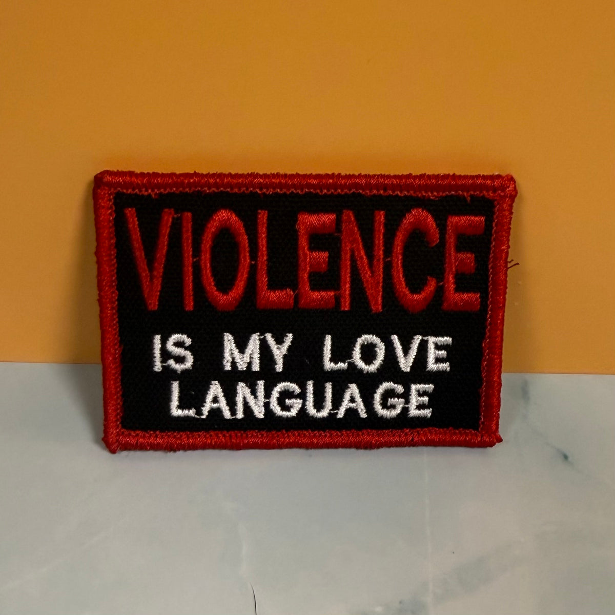 An embroidered rectangular patch with a red border, black background, and the text 'VIOLENCE IS MY LOVE LANGUAGE' in red and white lettering, placed against a yellow wall and light surface.