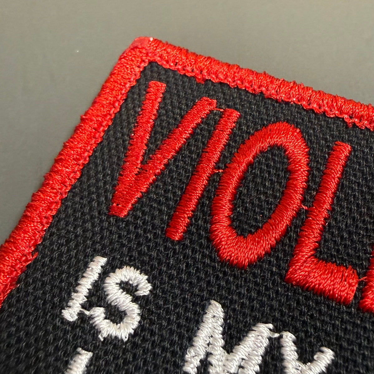 Close-up of an embroidered patch with a black background, red border and red lettering reading 'VIOLE', plus white lettering partially visible below.