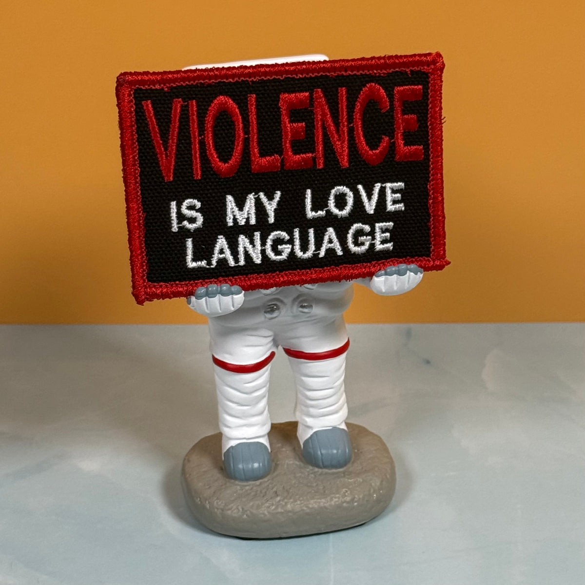 An embroidered patch with the text 'VIOLENCE IS MY LOVE LANGUAGE' in red and white on a black background, held by a small astronaut figurine with white spacesuit legs and a gray rock base, set against a solid orange background.