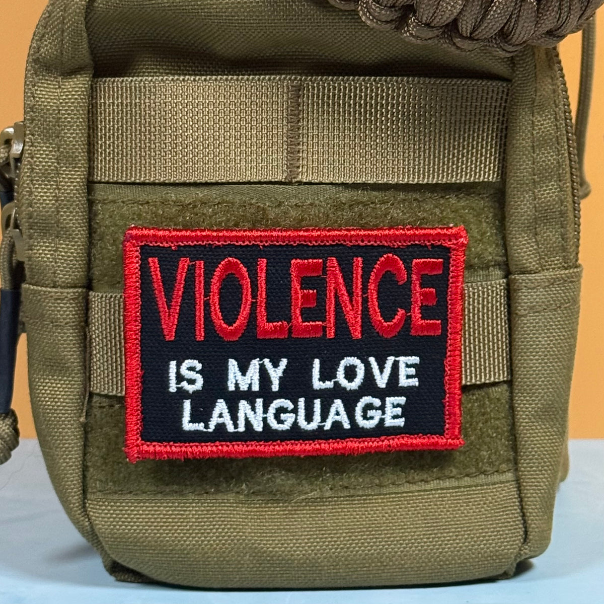 An olive green tactical pouch featuring a rectangular embroidered patch with the text 'VIOLENCE IS MY LOVE LANGUAGE' in red and white lettering on a black background.