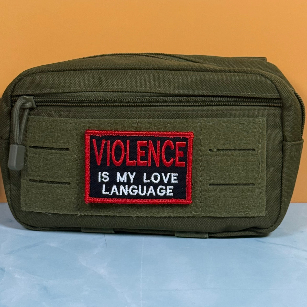 An olive green tactical pouch with a front zipper and a rectangular embroidered patch that reads 'VIOLENCE IS MY LOVE LANGUAGE' in red and white text on a black background, set against an orange and light blue backdrop.