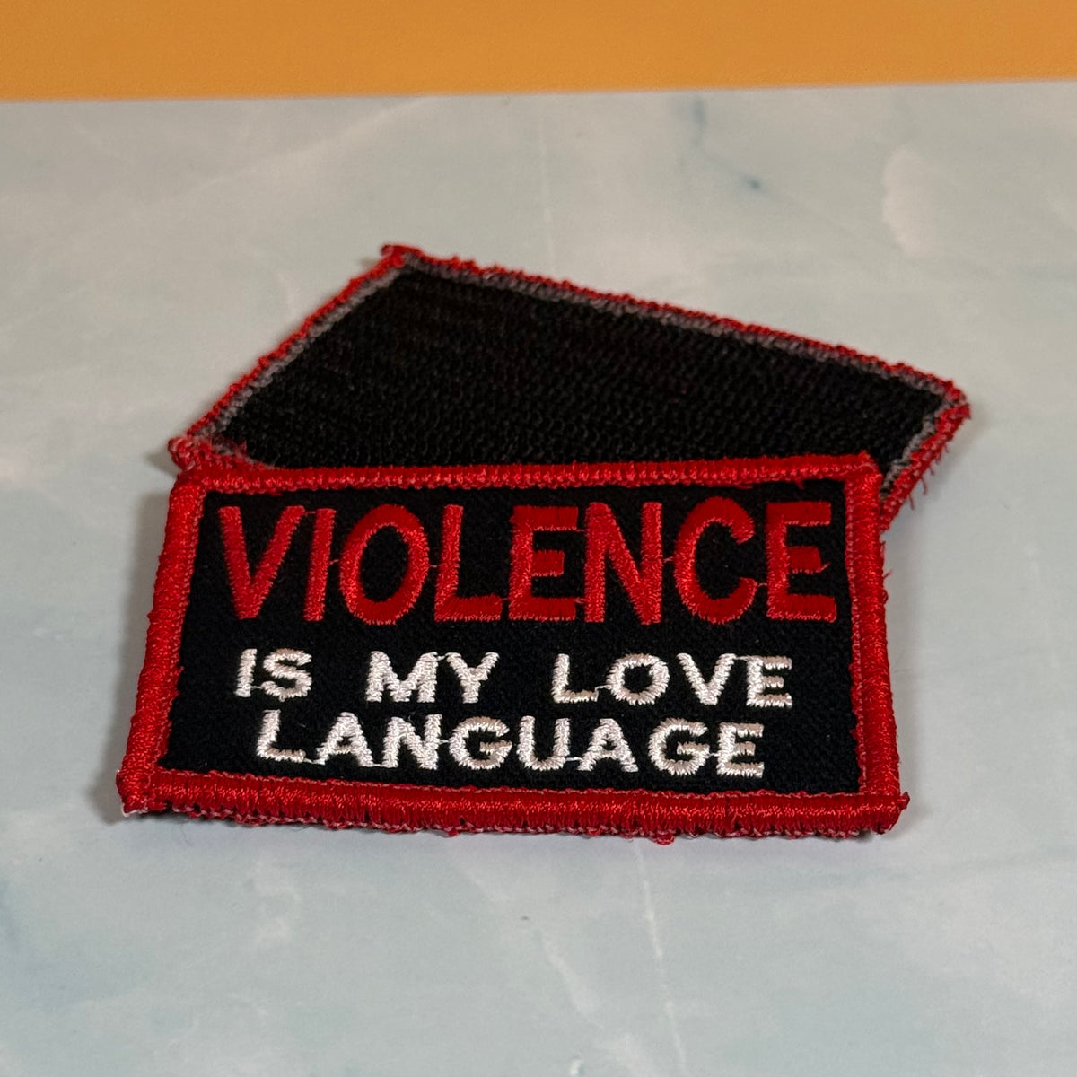 An embroidered rectangular patch with a black background, red border, and red and white text reading 'VIOLENCE IS MY LOVE LANGUAGE', shown on a light gray surface with an orange band at the top.