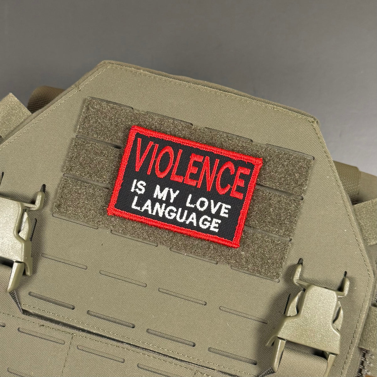 A rectangular embroidered patch with the text 'VIOLENCE IS MY LOVE LANGUAGE' in white on a black background with a red border, shown attached to a tan tactical vest or plate carrier.