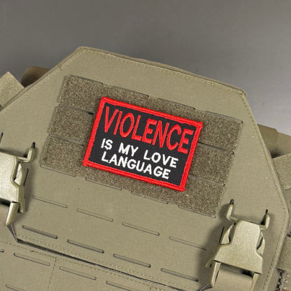 A rectangular embroidered patch with the text 'VIOLENCE IS MY LOVE LANGUAGE' in white on a black background with a red border, shown attached to a tan tactical vest or plate carrier.