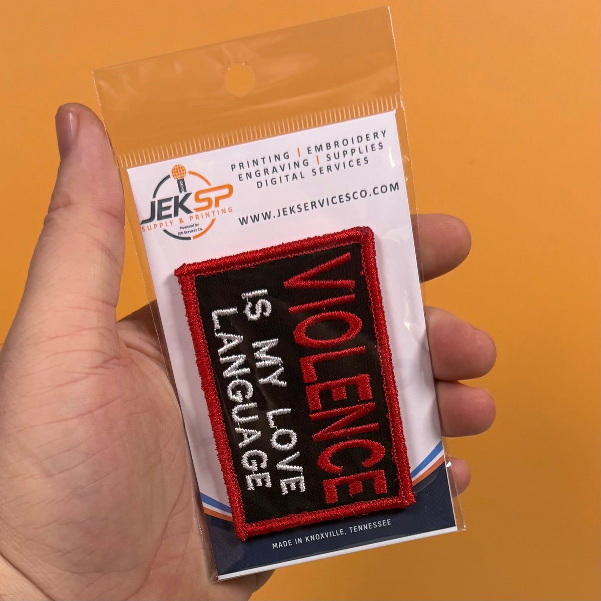 A hand holds a packaged embroidered patch that reads 'VIOLENCE IS MY LOVE LANGUAGE' in red and white on a black background. The packaging card shows the JEKSP Digital Embroidery logo and website, with a solid orange backdrop behind the hand.