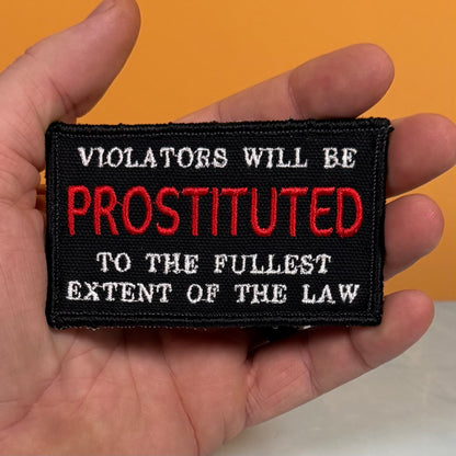 A hand holding a rectangular black embroidered patch with white and red text reading, 'VIOLATORS WILL BE PROSTITUTED TO THE FULLEST EXTENT OF THE LAW,' against an orange background.