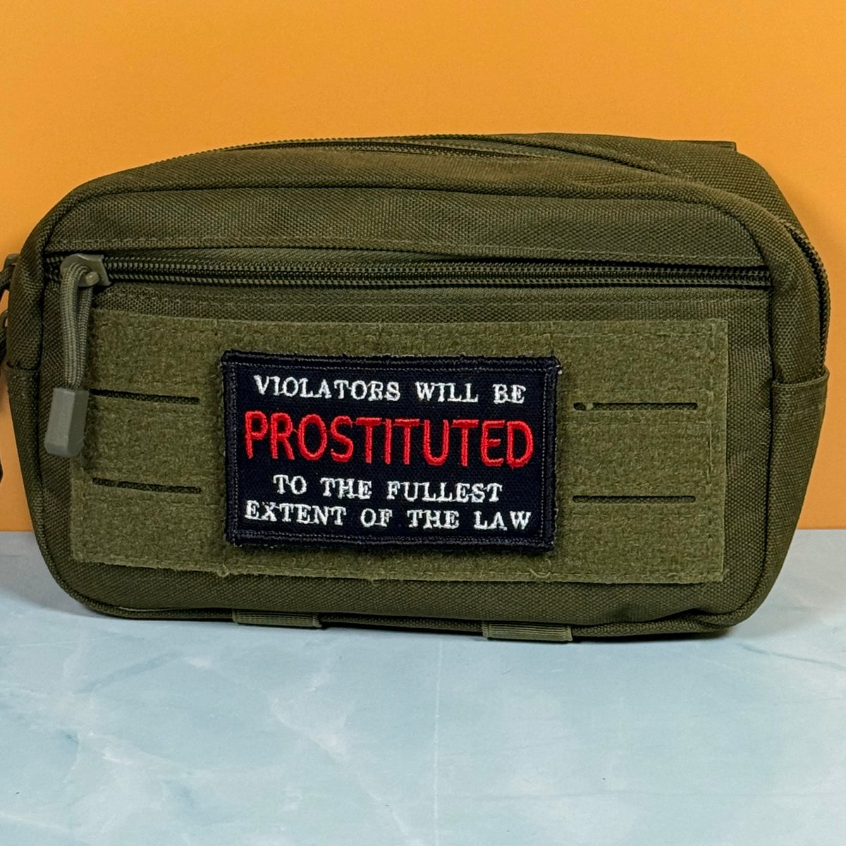 An olive green tactical pouch with a front zipper and a black embroidered patch featuring red and white text that reads, 'VIOLATORS WILL BE PROSTITUTED TO THE FULLEST EXTENT OF THE LAW.' The pouch is photographed against a solid orange background with a light blue surface below.