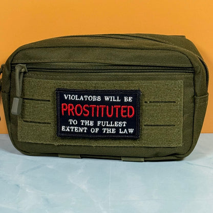 An olive green tactical pouch with a front zipper and a black embroidered patch featuring red and white text that reads, 'VIOLATORS WILL BE PROSTITUTED TO THE FULLEST EXTENT OF THE LAW.' The pouch is photographed against a solid orange background with a light blue surface below.