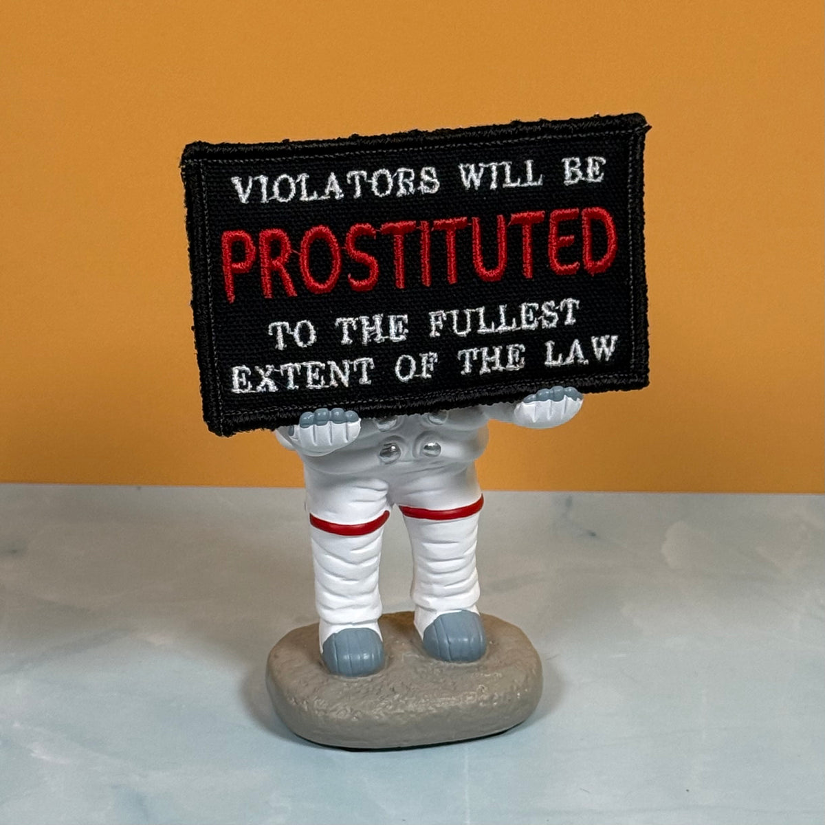 A novelty figurine of an astronaut holding a black embroidered sign that reads 'VIOLATORS WILL BE PROSTITUTED TO THE FULLEST EXTENT OF THE LAW' in white and red text, set against an orange background.