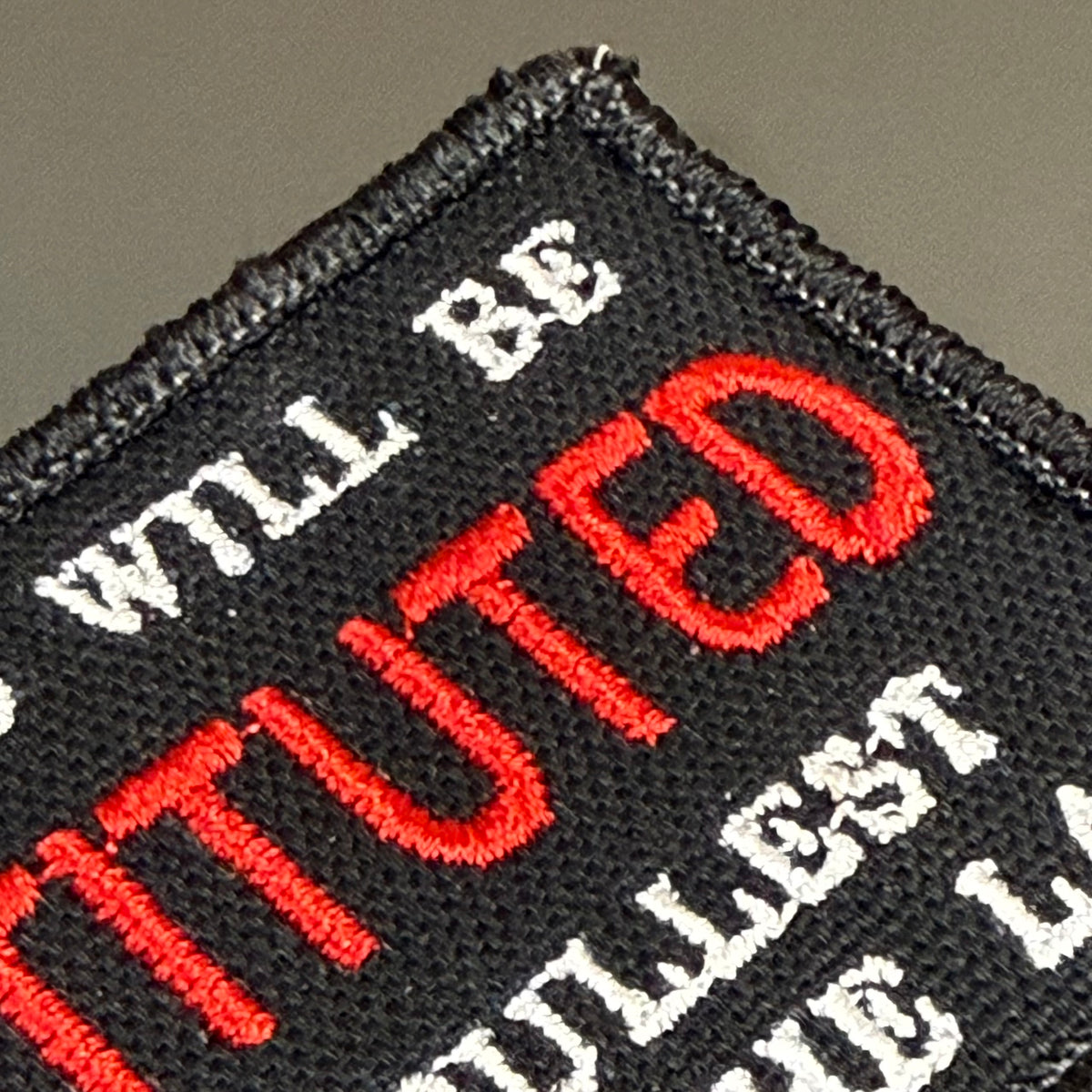 Close-up of a black embroidered patch with red and white text reading 'WILL BE INSTITUTED' on a dark gray background.