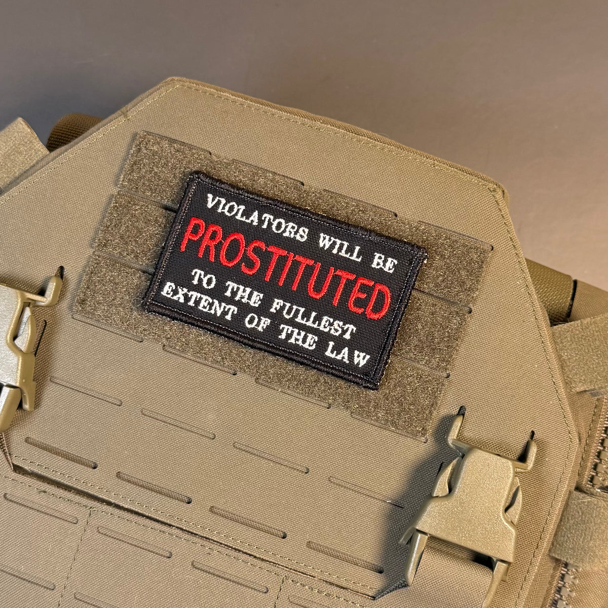 A tactical patch with the text 'VIOLATORS WILL BE PROSTITUTED TO THE FULLEST EXTENT OF THE LAW' in white and red lettering on a black background, mounted on a tan tactical vest.