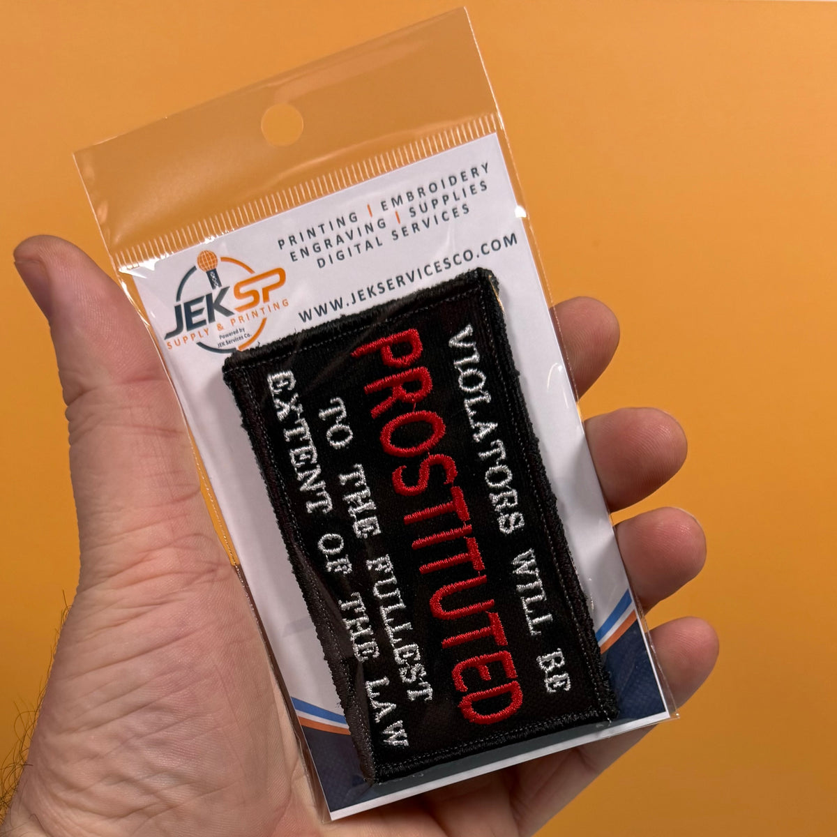 A hand holds a packaged embroidered patch against a solid orange background. The patch is black with red and white text reading 'PROSTITUTED' and 'VIOLATORS WILL BE TO THE FULLEST EXTENT OF THE LAW.