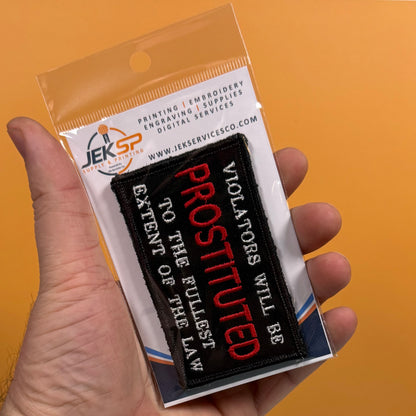 A hand holds a packaged embroidered patch against a solid orange background. The patch is black with red and white text reading 'PROSTITUTED' and 'VIOLATORS WILL BE TO THE FULLEST EXTENT OF THE LAW.
