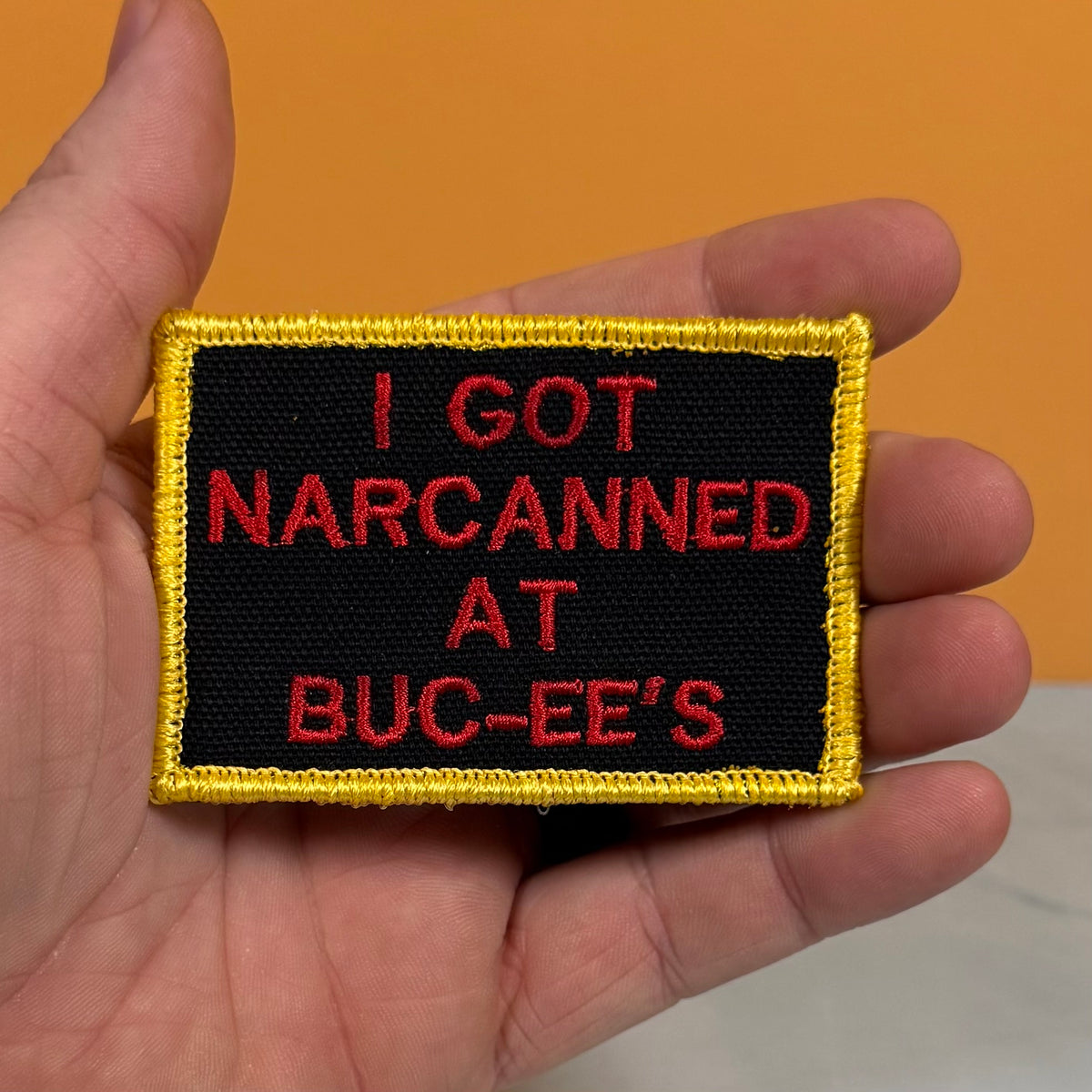 A hand holding a rectangular embroidered patch with a black background, red text reading 'I GOT NARCANED AT BUC-EE'S', and a yellow border against an orange background.