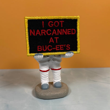 A small astronaut figurine holding a rectangular embroidered sign that reads 'I GOT NARCANED AT BUC-EE'S' in red lettering on a black background with a gold border. The figurine stands on a gray rock base against an orange wall and light gray surface.