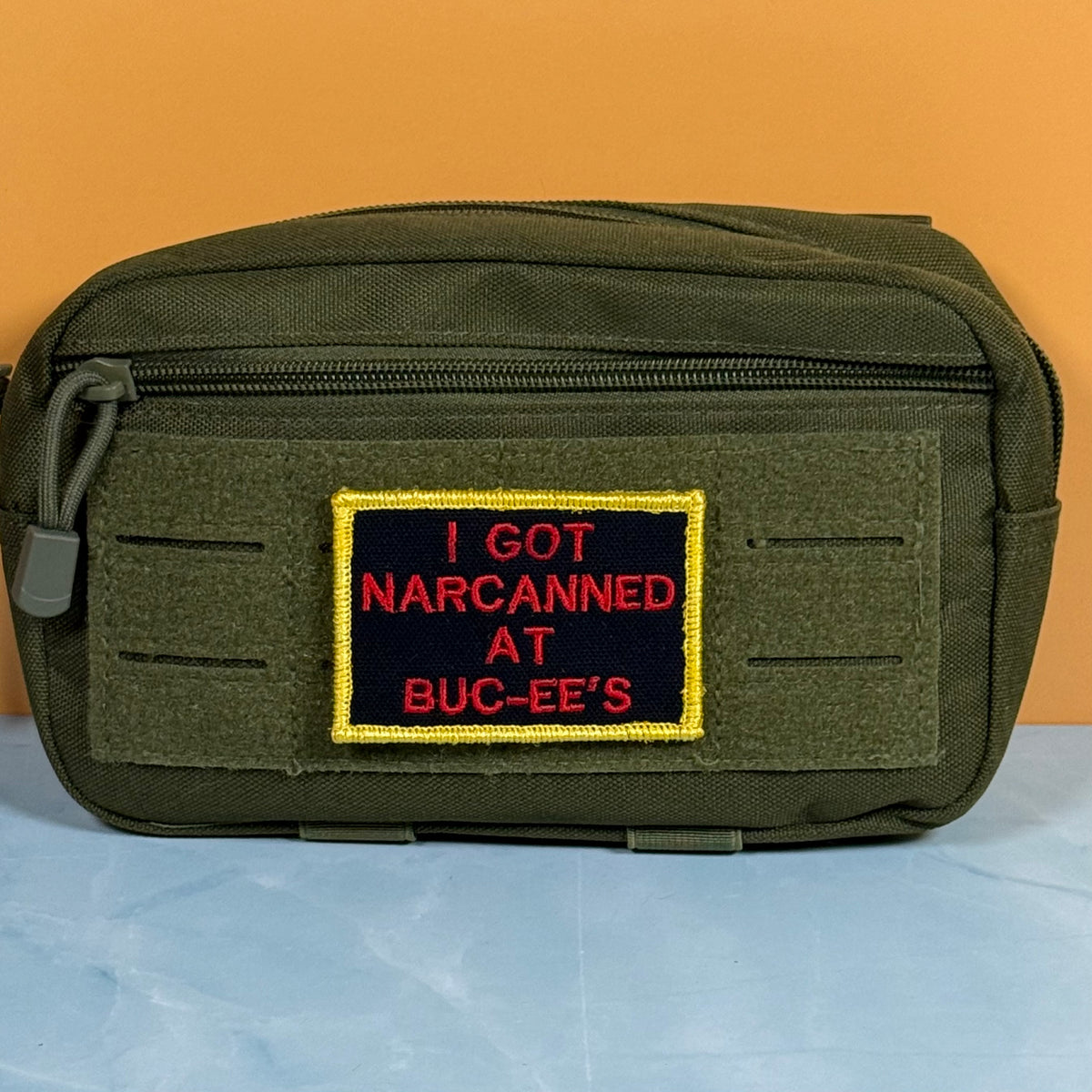 An olive green tactical pouch with a front zipper and a rectangular embroidered patch that reads 'I GOT NARCANNED AT BUC-EE'S' in red on a black background with a yellow border, set against an orange and light blue background.