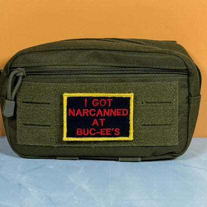An olive green tactical pouch with a front zipper and a rectangular embroidered patch that reads 'I GOT NARCANNED AT BUC-EE'S' in red on a black background with a yellow border, set against an orange and light blue background.